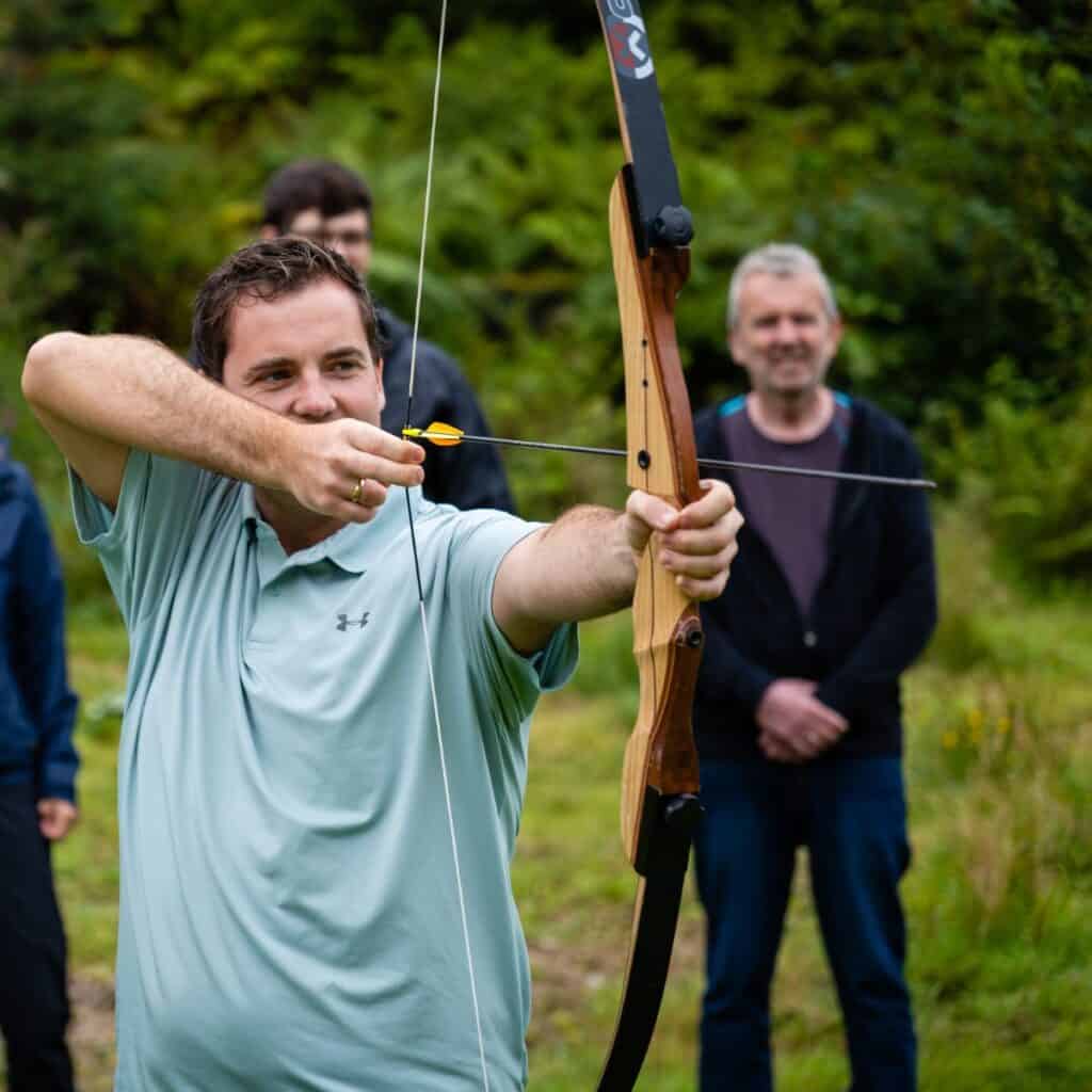 Skytter med bue på Sotra Villmarkssenter, omgitt av grønn natur og entusiastiske deltakere.
