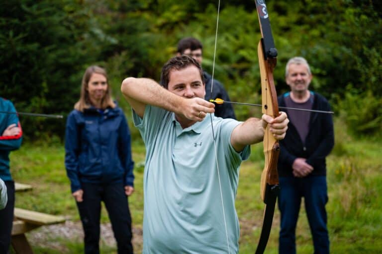 Mann som skyter med tradisjonell bue, omgitt av barn og voksne i naturen, ved Sotra Villmarkssenter, perfekt for friluftsliv.