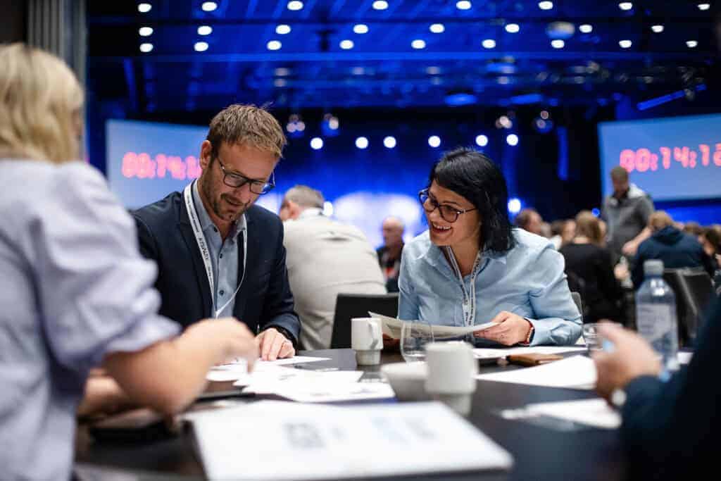 Bilde av mennesker som smiler og snakker under et konferansearrangement innenfor Sotra Villmarkssenter i Norge.